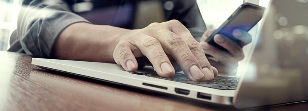 A person typing on a laptop with a cell phone